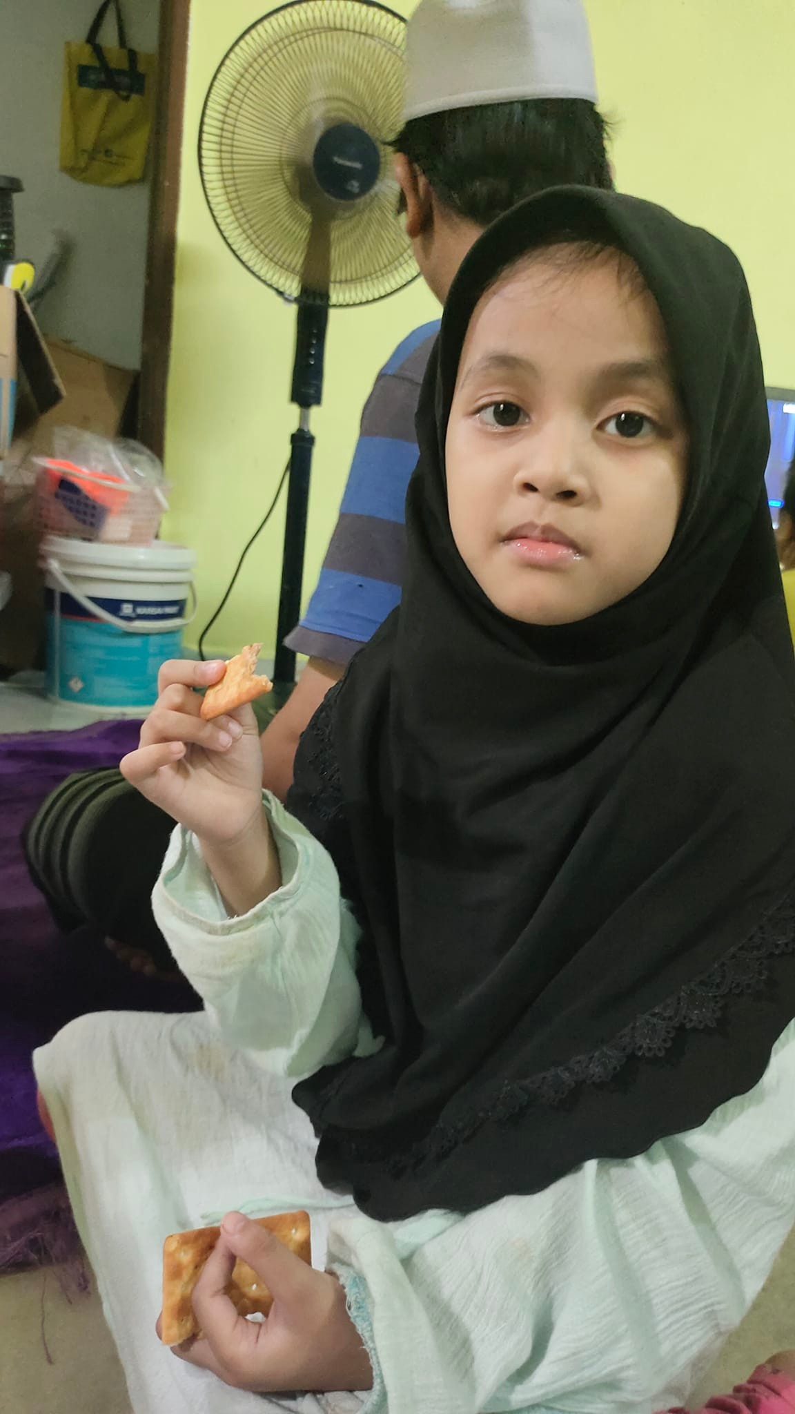 Ameera duduk tenang sambil memegang biskuit, disinari cahaya hangat. Di latar belakang, ayahnya sedang solat—suami saya—mewakili kehangatan keluarga dan ketabahan hidup bersama ibu yang menghidap bipolar.