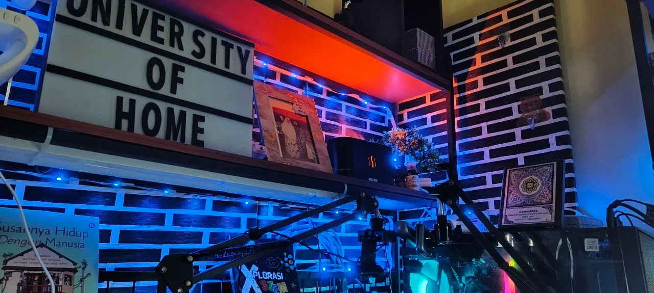 Sudut home studio dengan papan “University of Home”, lampu LED dan setup mikrofon di meja kerja
