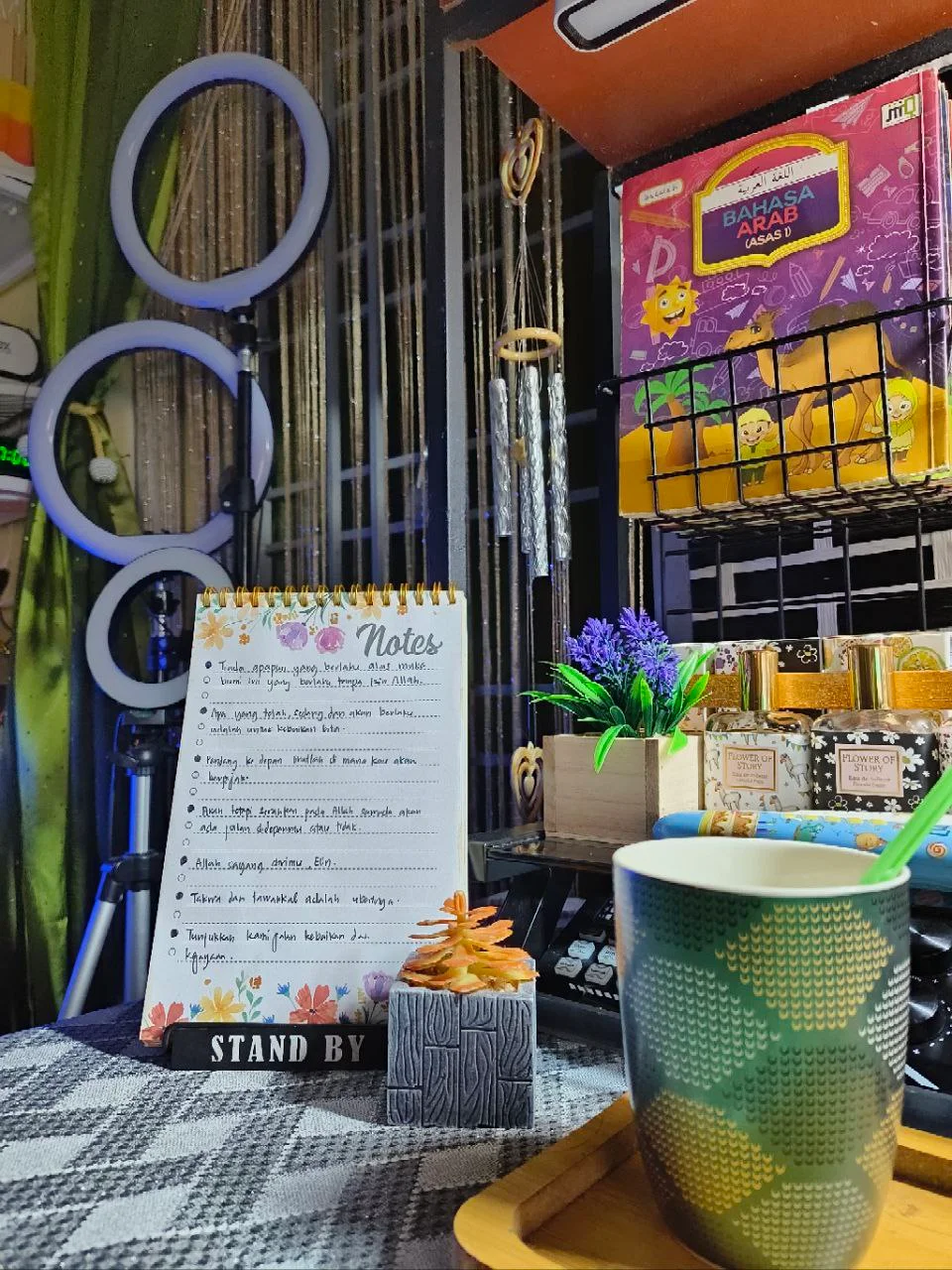 Sudut meja kerja dengan nota harian, cawan minuman, lampu ring light dan hiasan ruang kreatif di rumah